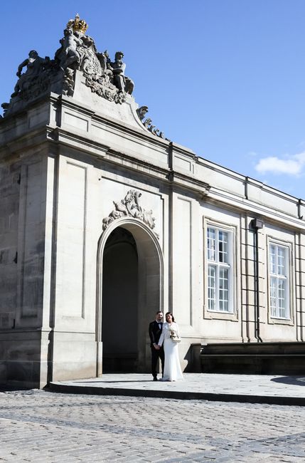 A bride and groom standing outdoors in front of an ornate, historic building with classical architecture, featuring large white stone walls, decorative sculptures atop the roof, and tall windows. The 