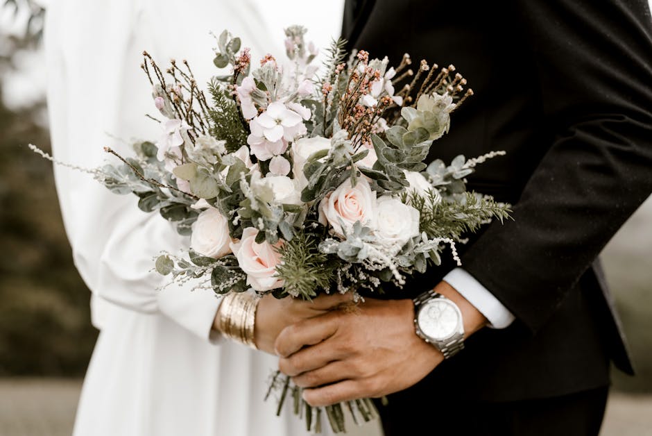 Close-up of a floral bouquet featuring soft pink roses, white anemones, and delicate sprigs of greenery, wrapped in light-colored tissue paper. The bouquet is held by a person wearing a white dress or