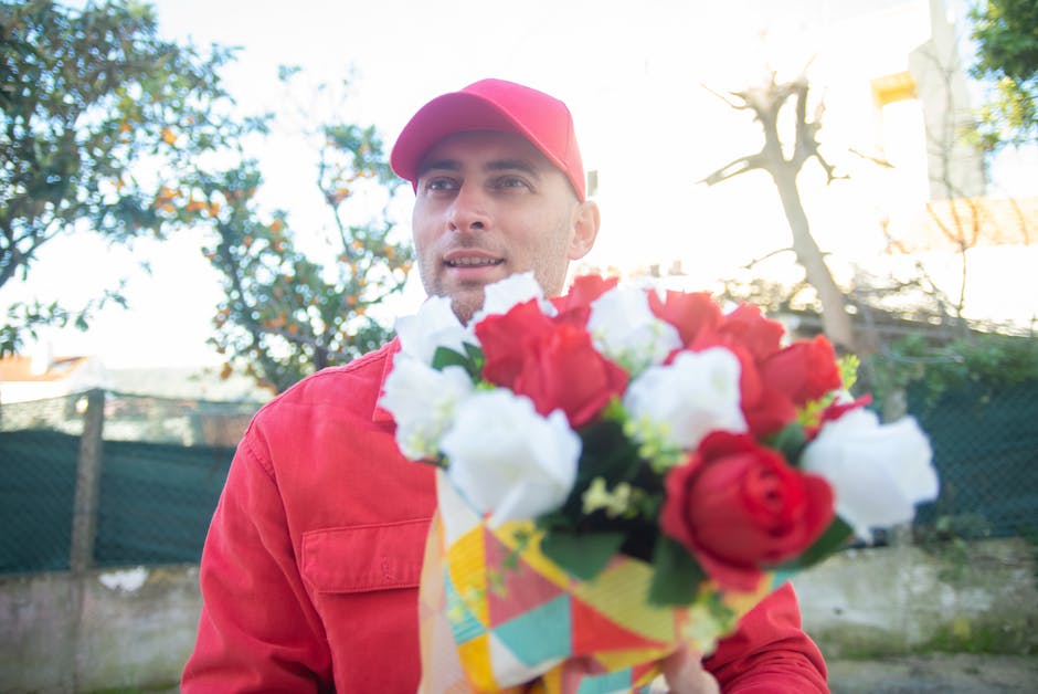 A man wearing a red jacket and matching red cap holds a vibrant floral bouquet featuring red and white roses, with some green foliage interspersed. He stands outdoors in a garden or park setting, with