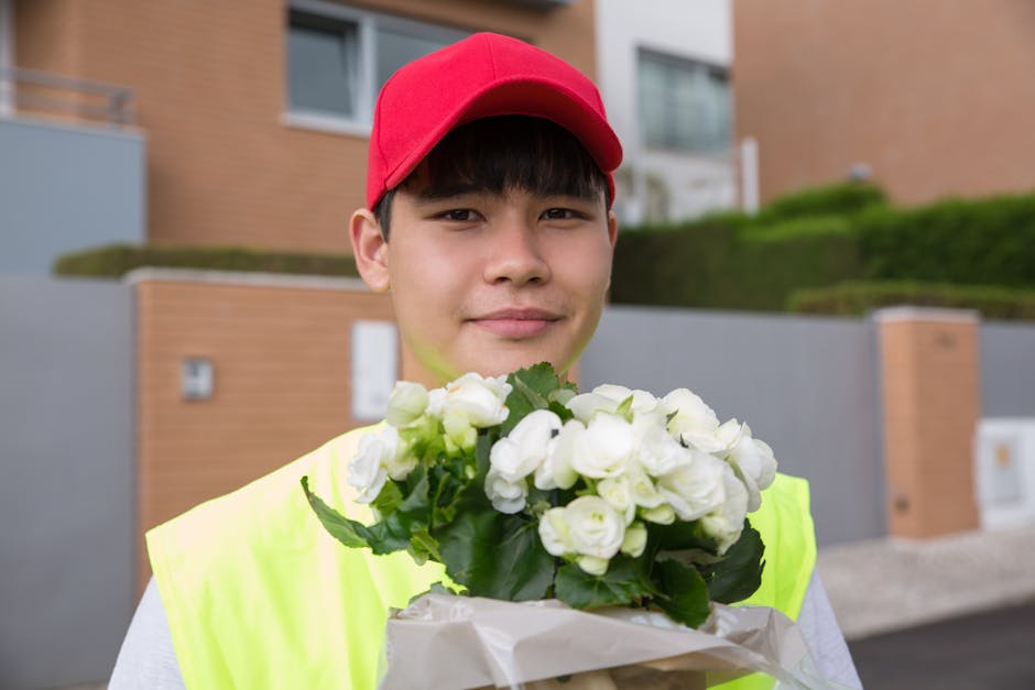 A young delivery person wearing a red cap and a neon yellow vest is outdoors, holding a bouquet of fresh white flowers wrapped in white paper. The flowers include white roses and freesias, with green 