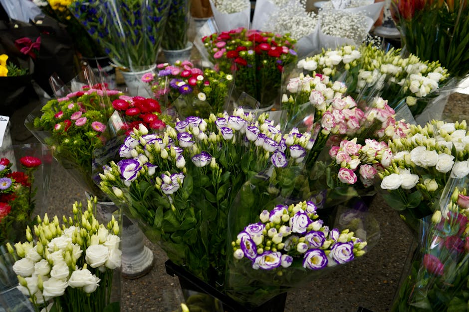 An assortment of fresh flowers displayed on a market table, including white eustoma, purple and white lisianthus, pink and white roses, and red and pink daisies, all arranged in clear plastic wrapping